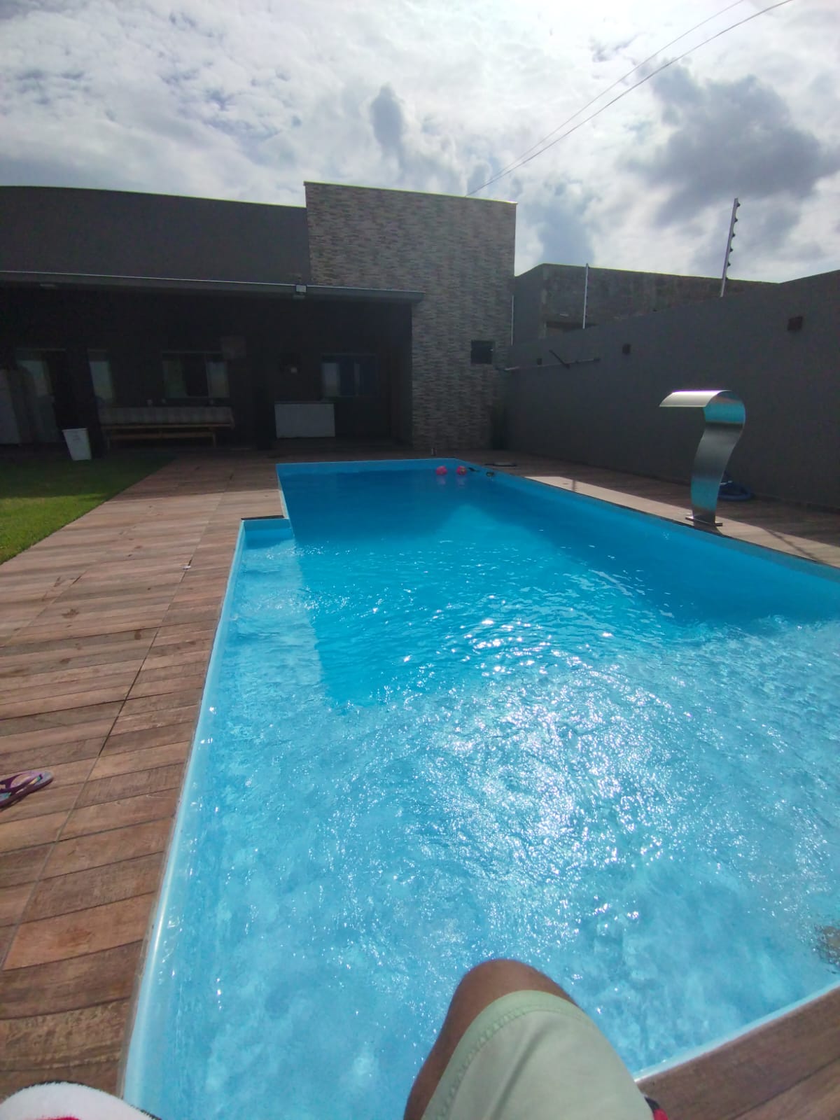 A large swimming pool is framed by wooden decking, reflecting sunlight on its clear surface. The pool area is surrounded by a green lawn, with a distant view of a covered patio featuring seating. The scene is complemented by a cloudy sky and modern architectural elements.