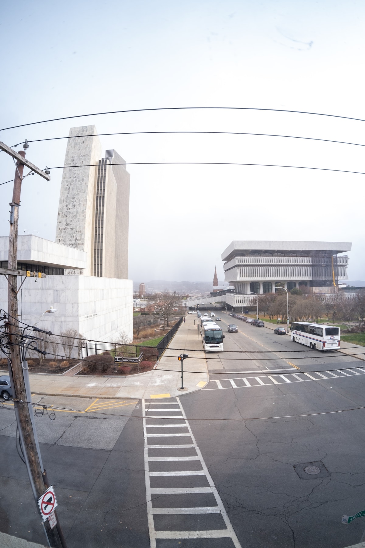 A cityscape view features a blend of modern architecture, including a tall concrete building and a large structure with a distinctive wide roof. A wide street lined with light poles and traffic signs runs through the foreground, with vehicles visible in motion.