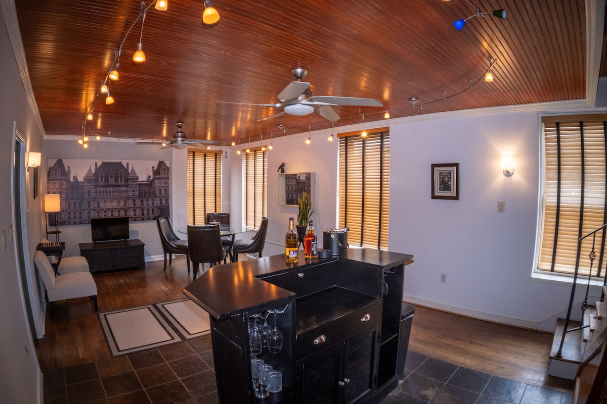 An open living space displays a blend of modern and classic design, featuring a dark kitchen island with bar stools. Warm lighting highlights the wood-paneled ceiling, while a comfortable seating area is visible in the background, alongside large windows with wooden blinds.
