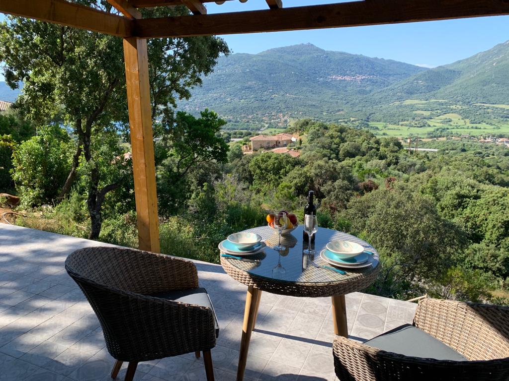 A dining area features a round table set for two, with plates and glassware arranged neatly. Two woven chairs frame the table. The outdoor space is surrounded by lush greenery, overlooking a picturesque valley and distant mountains under clear skies.
