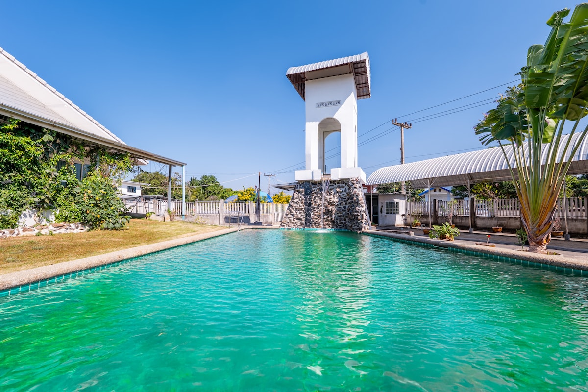 A large swimming pool is displayed, featuring clear turquoise water. A stone waterfall cascades elegantly at one end, framed by lush greenery. The surrounding area has a sun-drenched deck, providing an inviting space for relaxation.