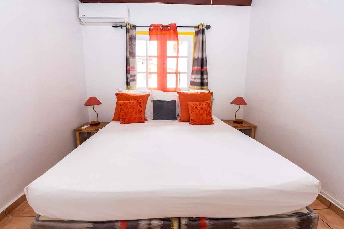 A spacious sleeping area is presented with a large bed topped with a white mattress and adorned with orange decorative pillows. Small bedside tables with lamps are positioned on either side. Light streams in through a window dressed with sheer and patterned curtains.