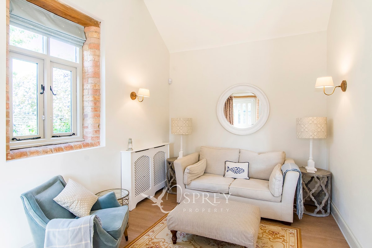 A bright seating area features a spacious light-colored sofa and a cozy armchair, complemented by decorative table lamps. A rounded mirror enhances the wall above the sofa, while a window allows natural light to fill the room.