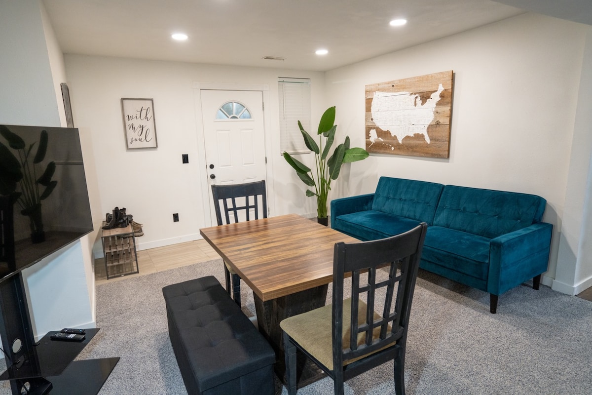 A modern living area features a comfortable blue sofa and a wooden dining table surrounded by chairs. A potted plant adds a touch of greenery. A map of the United States is mounted on the wall, and natural light filters through the front door.