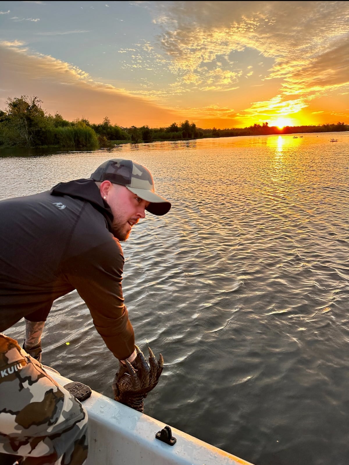 The image captures a person extending their hand towards the water, while a vibrant sunset casts warm hues across the sky and reflects on the surface. Silhouettes of trees line the horizon, enhancing the natural beauty of the tranquil setting.