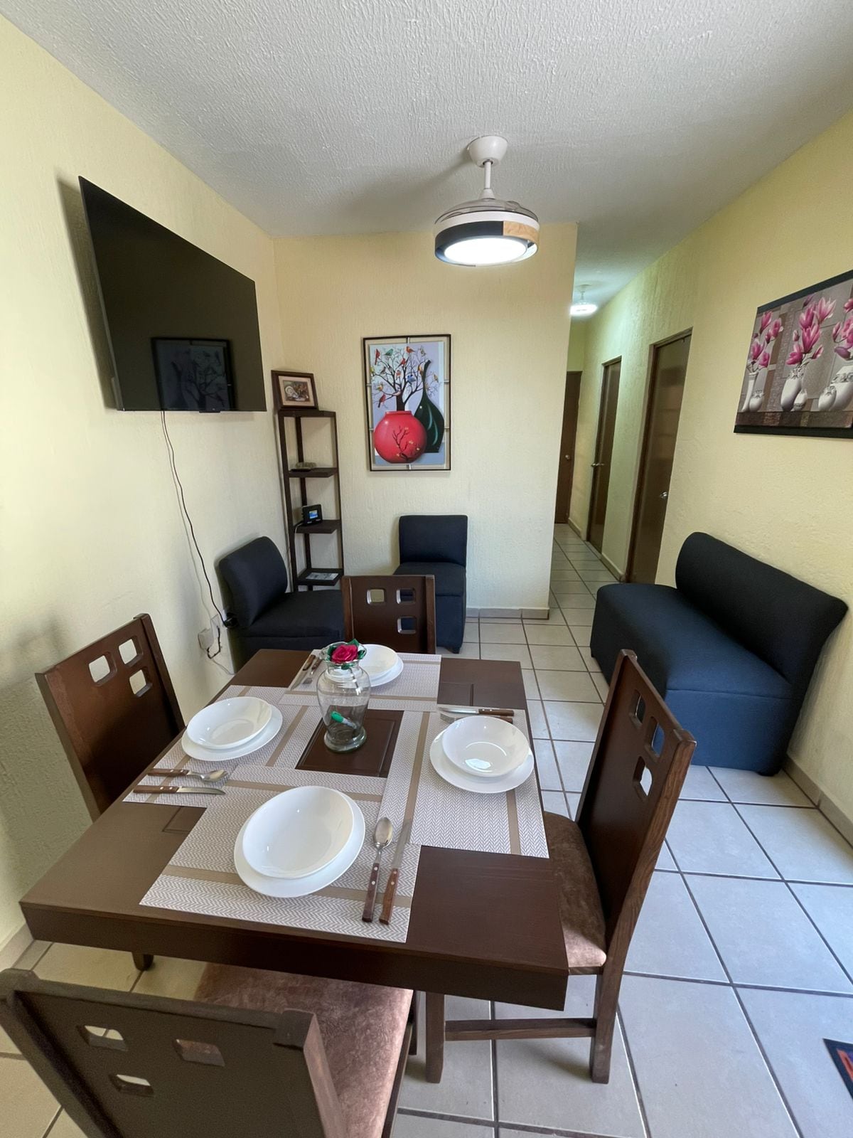A dining area features a wooden table set for four, accompanied by dark upholstered chairs. A television is mounted on the wall above a narrow shelf. Light-colored walls are complemented by framed artwork, and a cozy seating area is visible in the background.