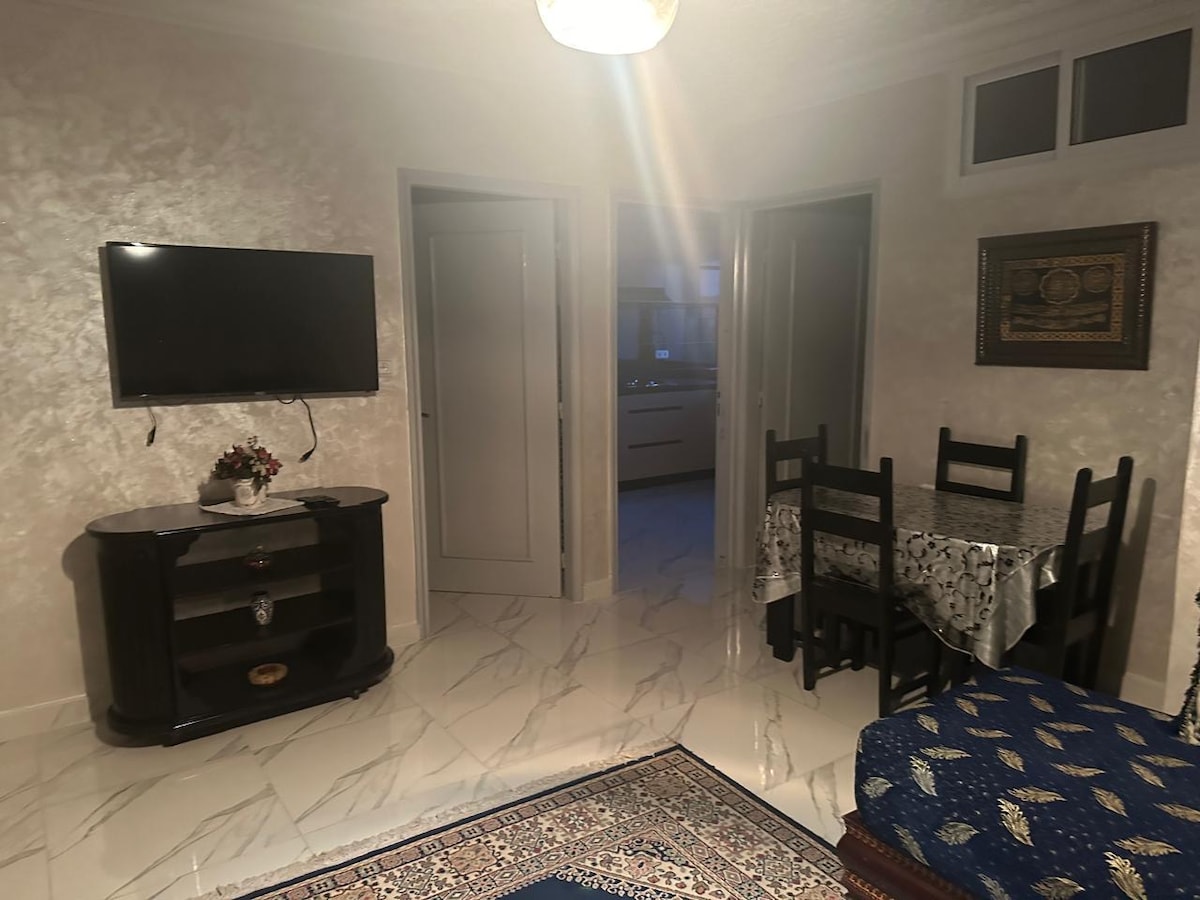 A bright living area features a television mounted on the wall above a dark wooden console. A dining table with four chairs is positioned near a doorway leading to the kitchen, while decorative elements are visible throughout the space.