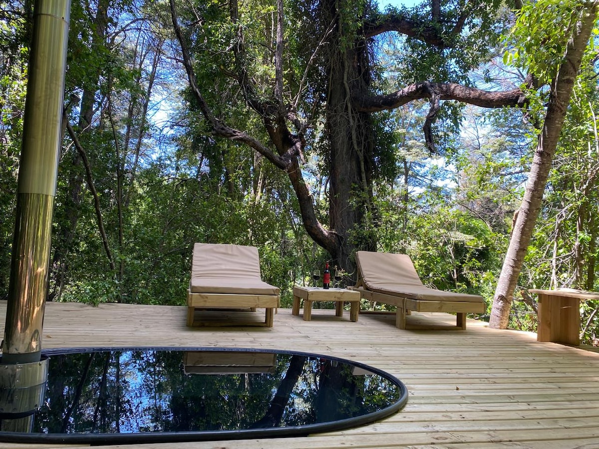 A wooden terrace is surrounded by lush greenery, featuring two lounge chairs positioned beside a dark circular hot tub. A small wooden table accompanies the chairs, creating a serene outdoor relaxation area amid the forest.