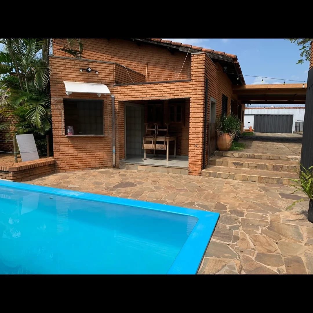 A bright outdoor area features a refreshing pool with a blue edge, surrounded by a stone patio. A brick house is visible in the background, showcasing large windows and a shaded seating area with wooden chairs. Lush greenery adds to the tranquil setting.
