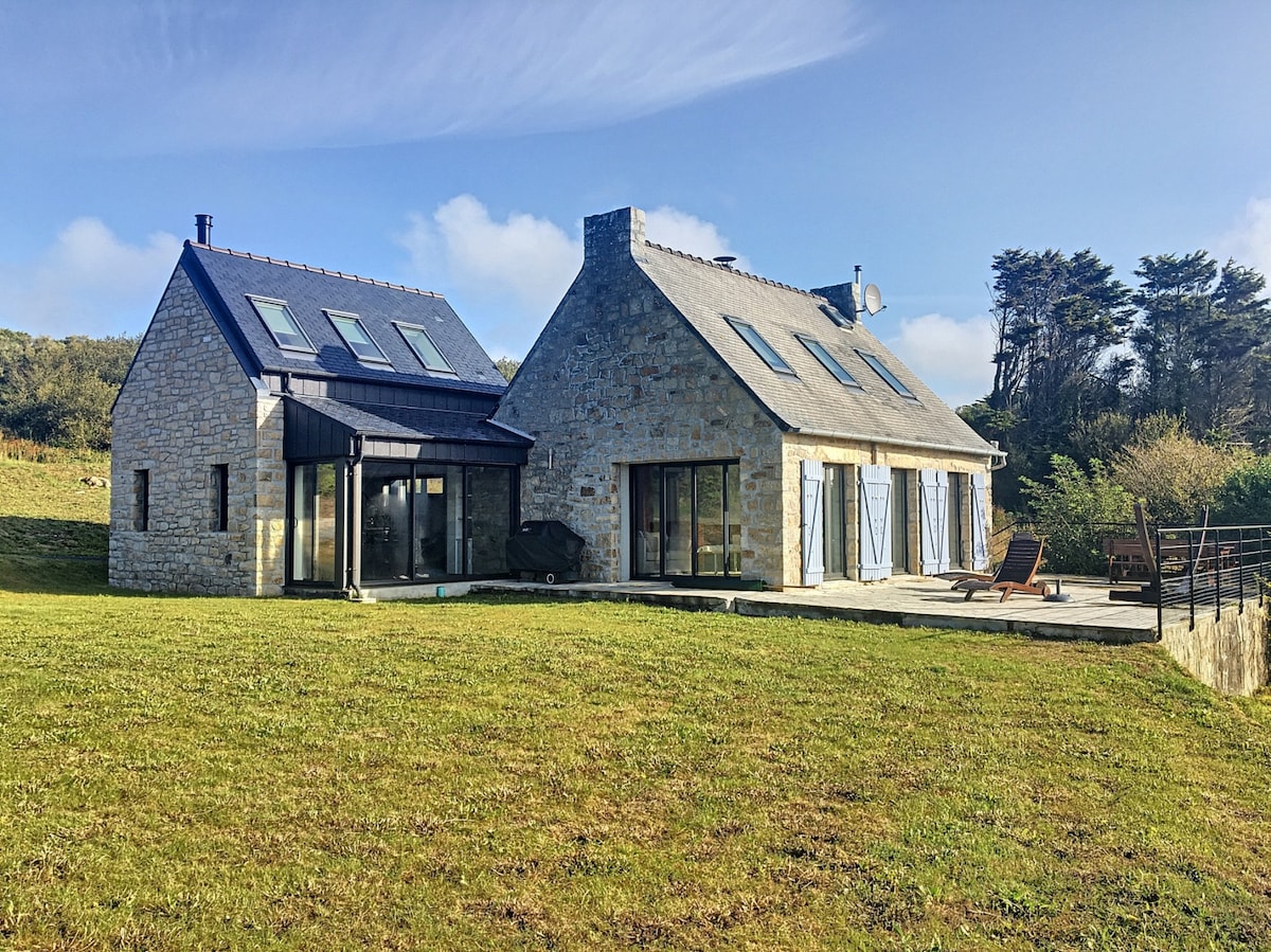 The stone-faced house is set against a backdrop of lush greenery, featuring a modern design. Large glass windows reflect natural light, and outdoor lounge chairs are arranged on the terrace. The surrounding landscape extends to rolling hills and trees, enhancing the serene atmosphere.