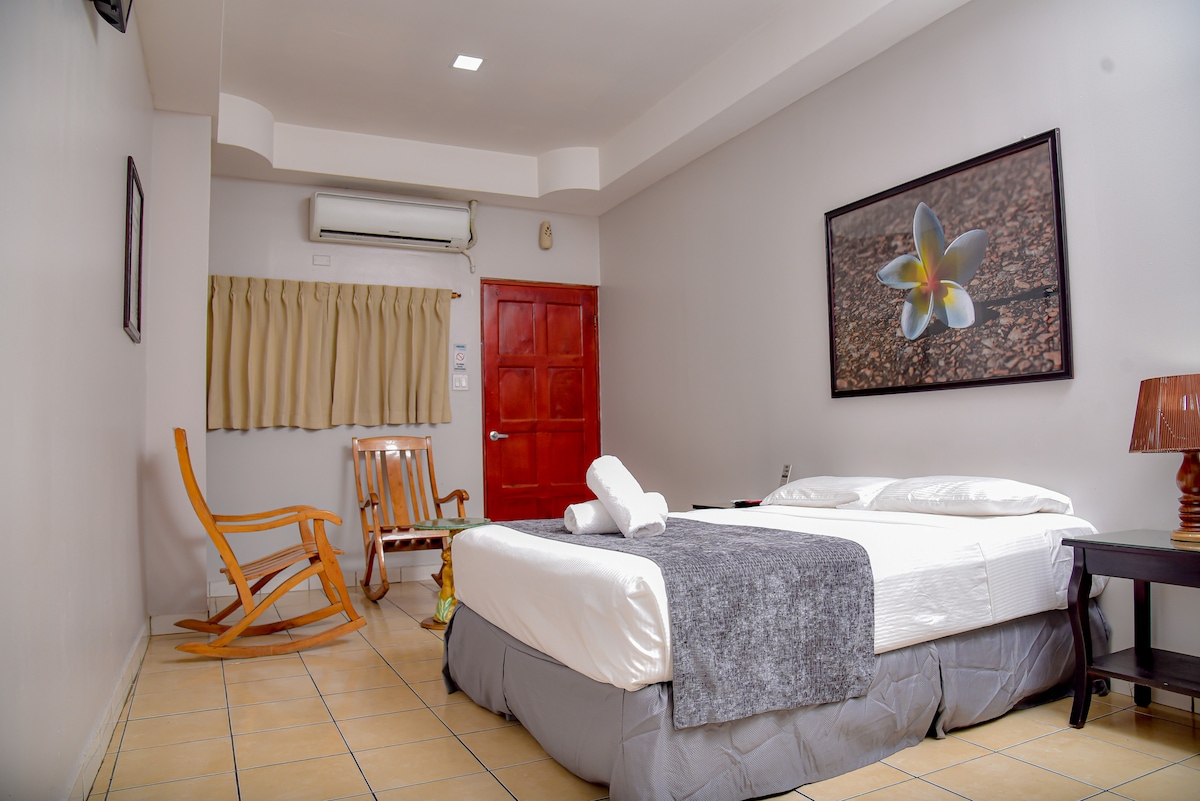 A well-lit room features a spacious bed with neatly arranged linens. A rocking chair is positioned beside the bed. A decorative artwork depicting a flower hangs on the wall. Natural light filters through a window covered by light curtains, and an air conditioning unit is mounted above.