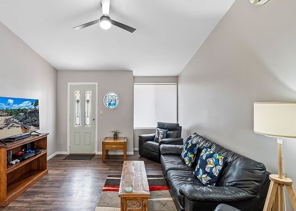 A light-filled living area features a leather lounge and recliner chairs, positioned around a wooden coffee table. A television is situated on a low shelf, and a round mirror reflects natural light near the entry door. A classic ceiling fan adds to the room's comfort.