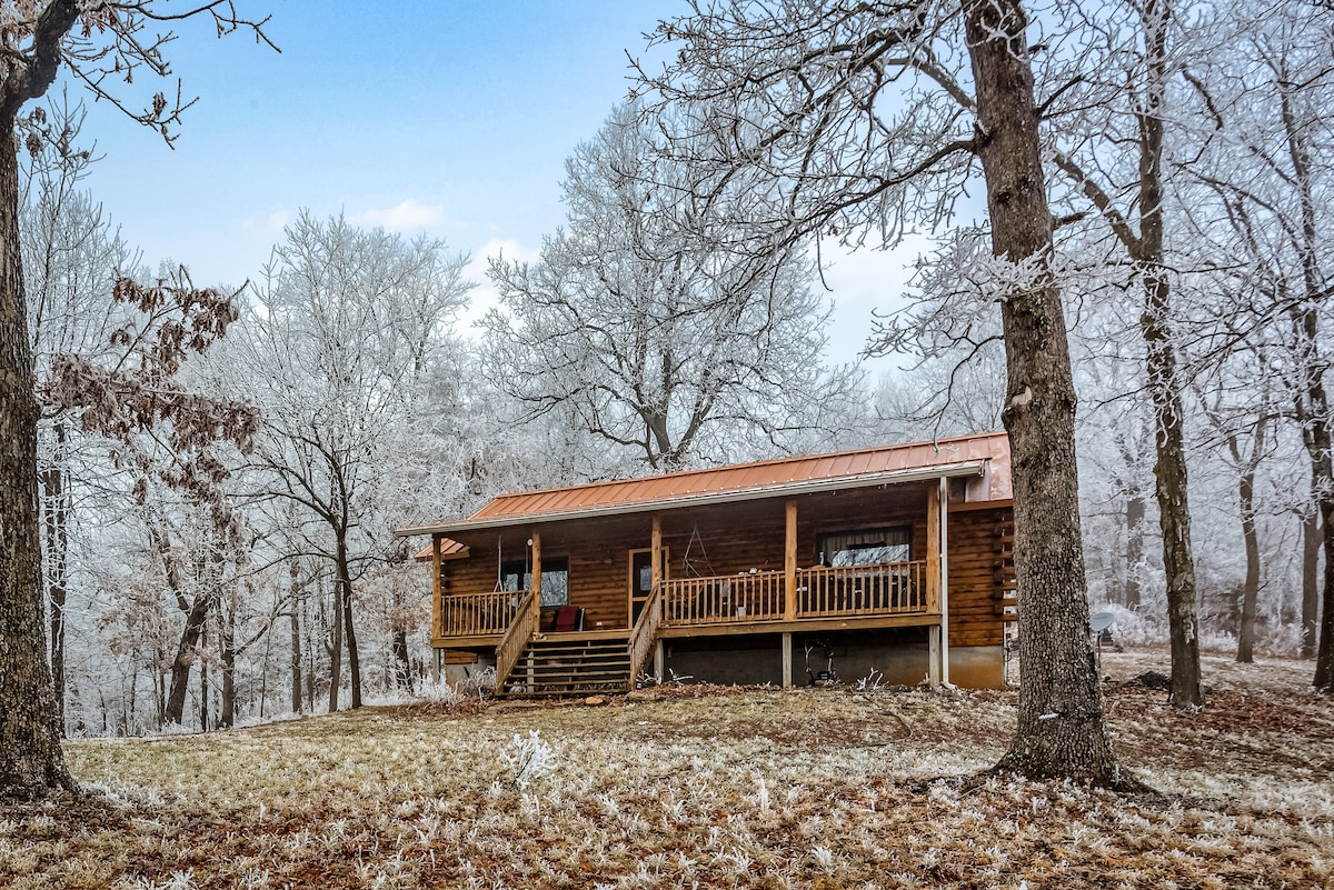 A log cabin is showcased amidst a serene winter landscape, surrounded by frost-covered trees. A large front porch is visible, equipped with a set of chairs, while the cabin's wooden structure exudes rustic charm against a clear blue sky.
