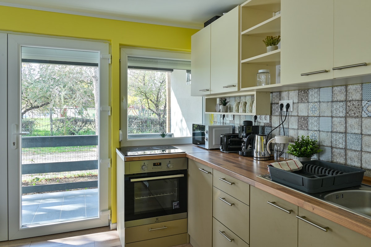 A modern kitchen featuring light-colored cabinetry, a wooden countertop, and stainless-steel appliances. Natural light fills the space through large sliding doors, which open to a view of the garden. A stylish backsplash enhances the overall design, contributing to a functional cooking area.