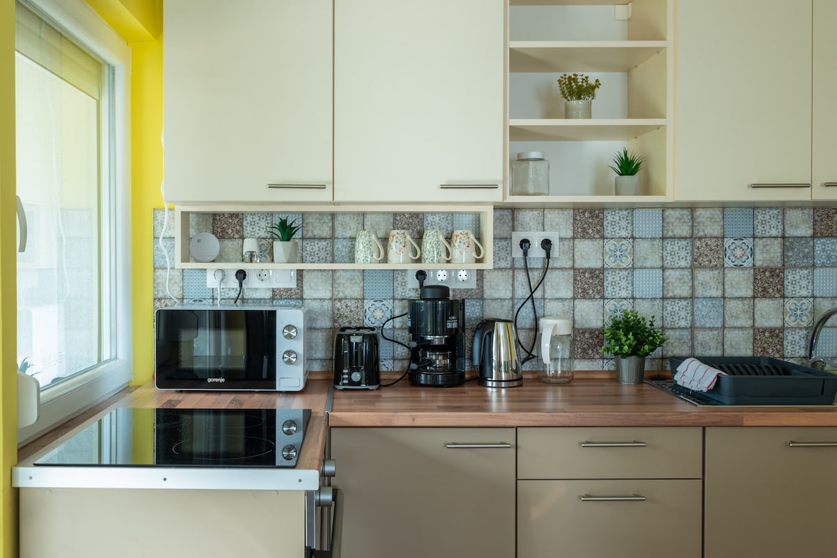 A modern kitchen is equipped with a microwave, coffee maker, and kettle on a wooden countertop. The beige cabinets are paired with a textured tile backsplash featuring diverse patterns. Natural light enters through a window, enhancing the clean and organized space.