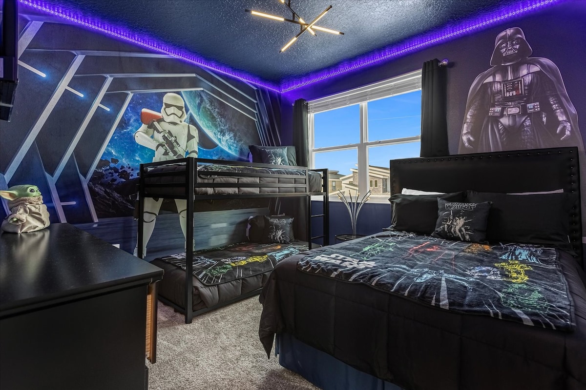 A themed bedroom features dark walls adorned with Star Wars graphics, including a prominent Darth Vader image. A bunk bed occupies one corner, covered with bedding featuring colorful space designs. A window allows natural light to fill the room, complemented by purple LED lights along the ceiling.