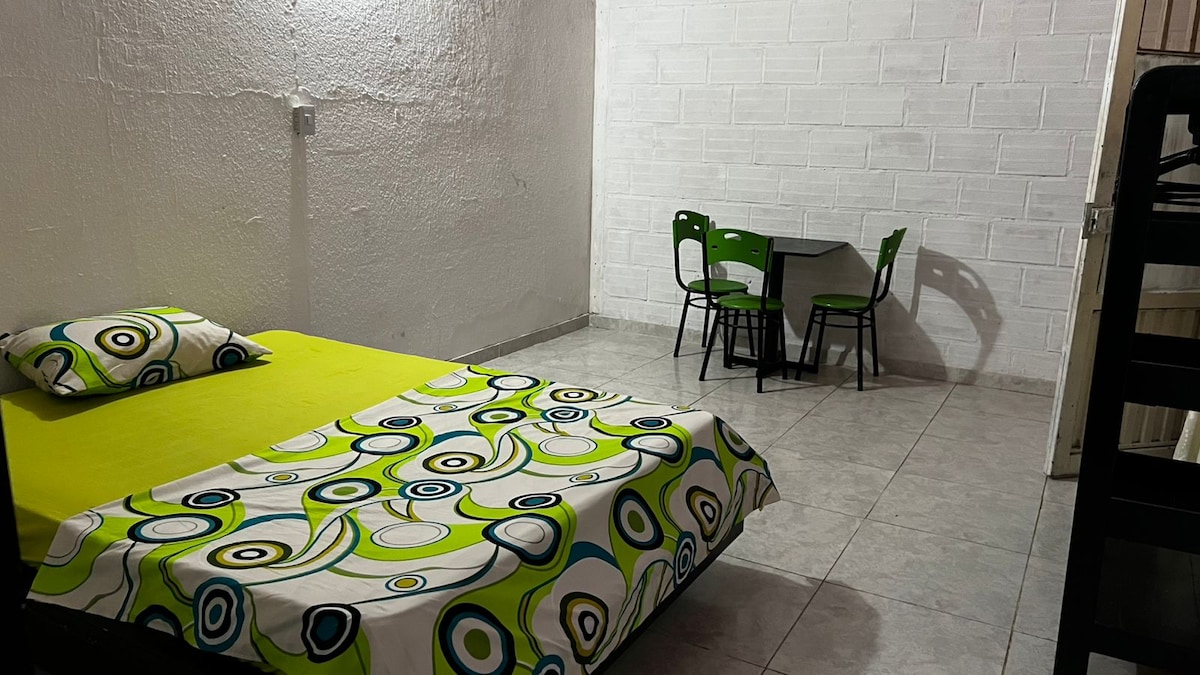 A spacious sleeping area features a bed covered with a vibrant, patterned blanket. A small dining table with two chairs is positioned nearby, set against a simple, white brick wall. The flooring is tiled, providing a clean and functional aesthetic.