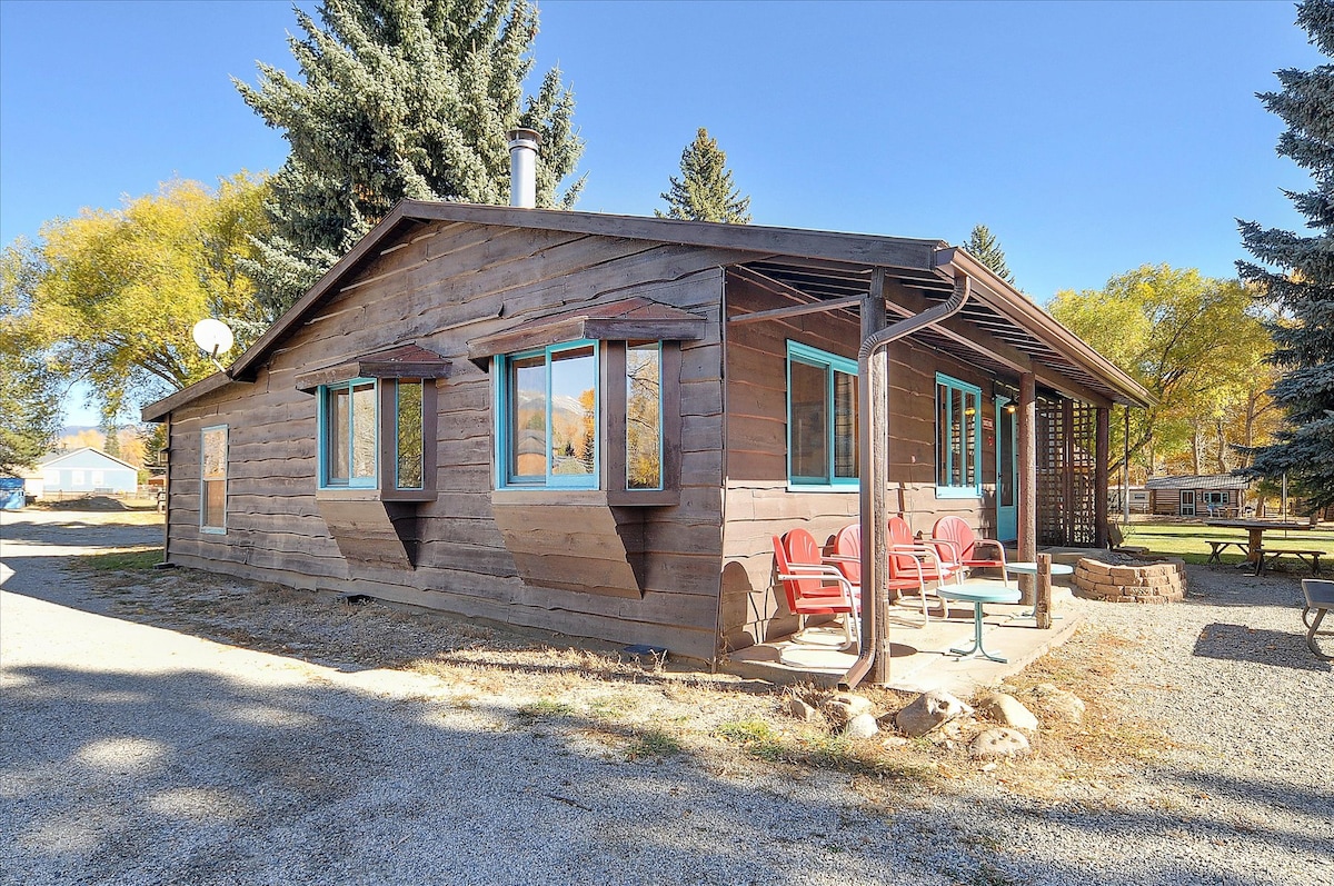 A wooden cottage exterior is visible, featuring large windows and brown siding. Outdoor seating is arranged on the front patio, with red chairs positioned around a small table. Nearby trees provide shade, complementing the open landscape.