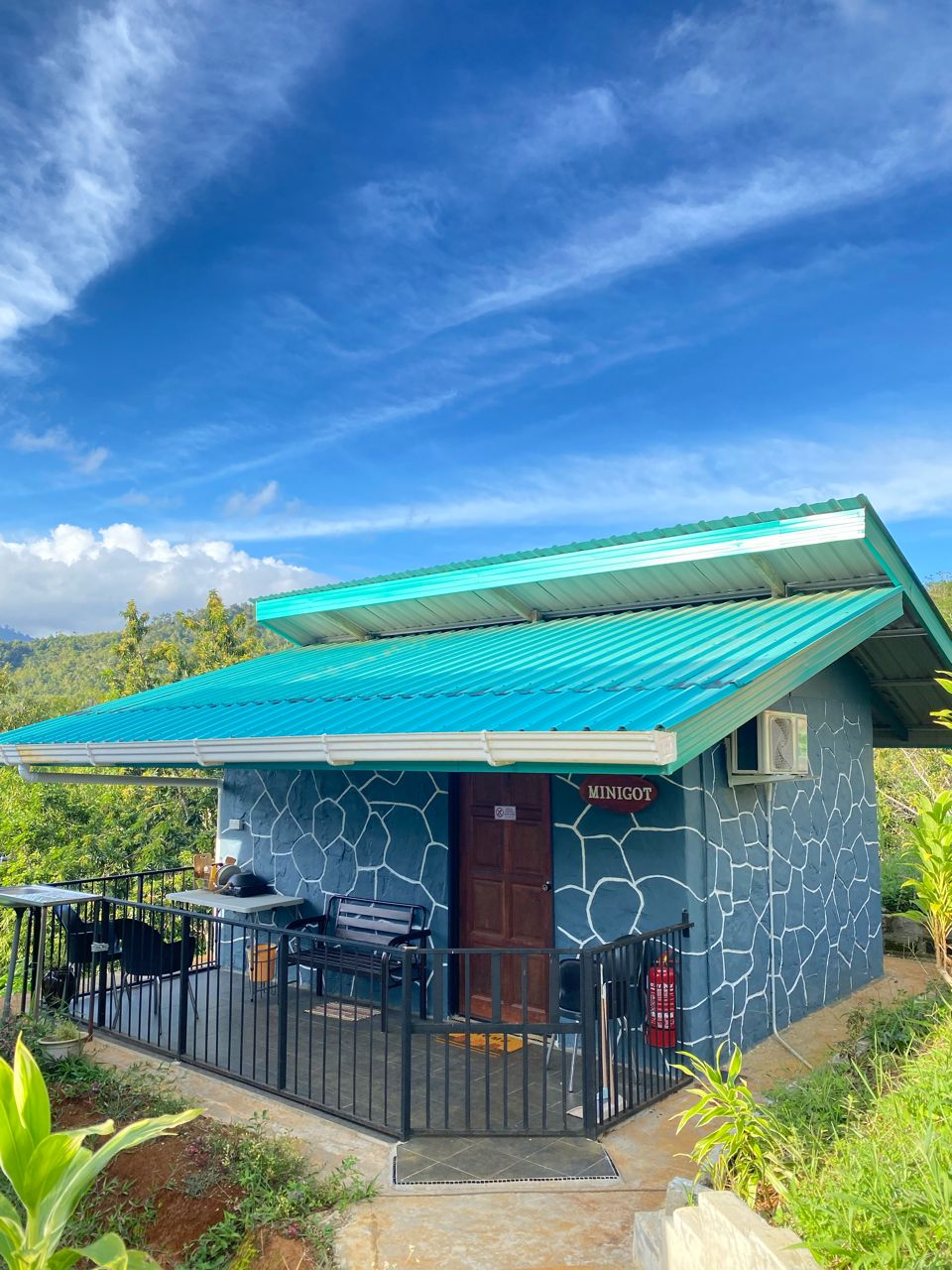 A charming cabin with a turquoise metal roof is framed by scenic greenery. The stone wall design adds character to the exterior. A covered porch area is furnished with black chairs, inviting relaxation in a tranquil environment.