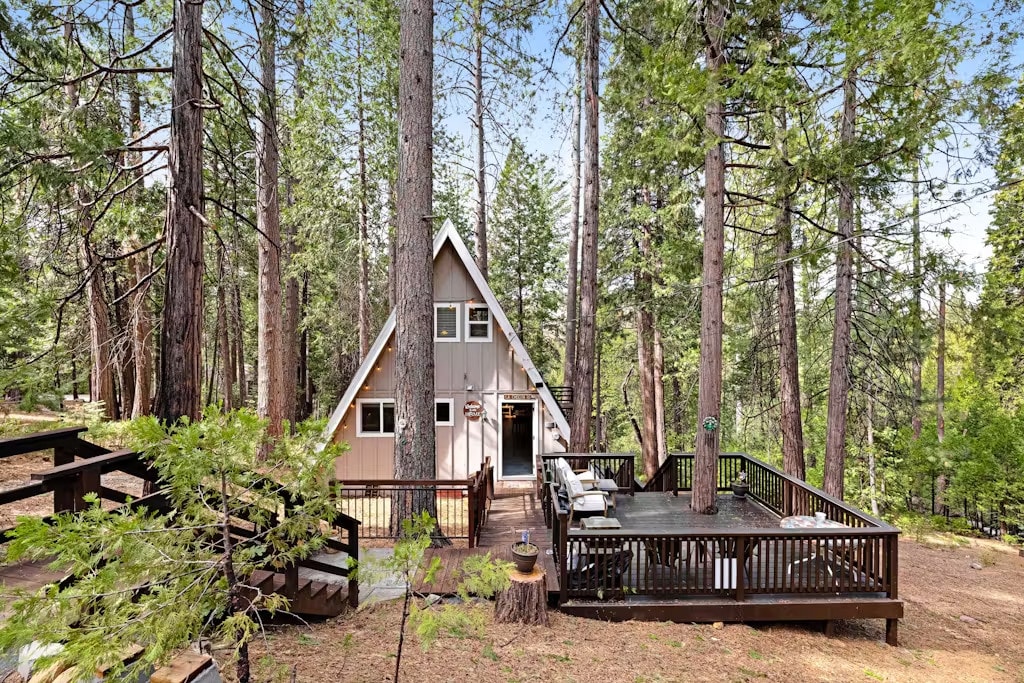 The A-frame cabin is surrounded by tall trees, showcasing a natural setting. A deck is visible in front of the cabin, equipped with seating. The main entrance is highlighted by large windows, and the roof features a distinctive triangular shape characteristic of A-frame designs.