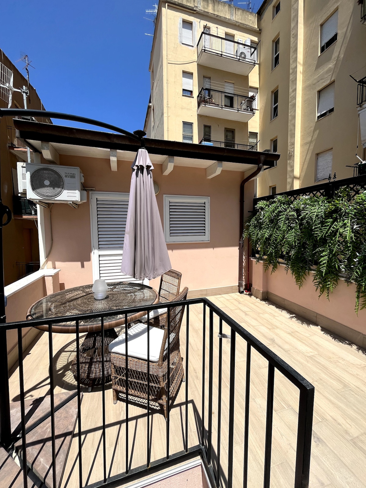 A private terrace features a round table with matching chairs, providing a comfortable outdoor seating area. A large umbrella offers shade, while a lush green plant hangs from the wall, adding a touch of nature. The space is illuminated by natural sunlight from a clear blue sky.