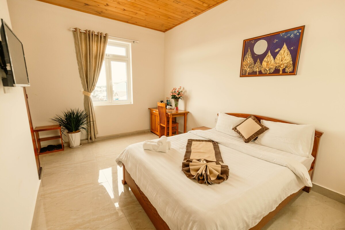 A well-arranged guest room features a spacious bed with neutral linens, complemented by decorative pillows. A window allows natural light to brighten the space, while a small table and chair sit nearby. A potted plant adds a touch of greenery, enhancing the serene atmosphere.