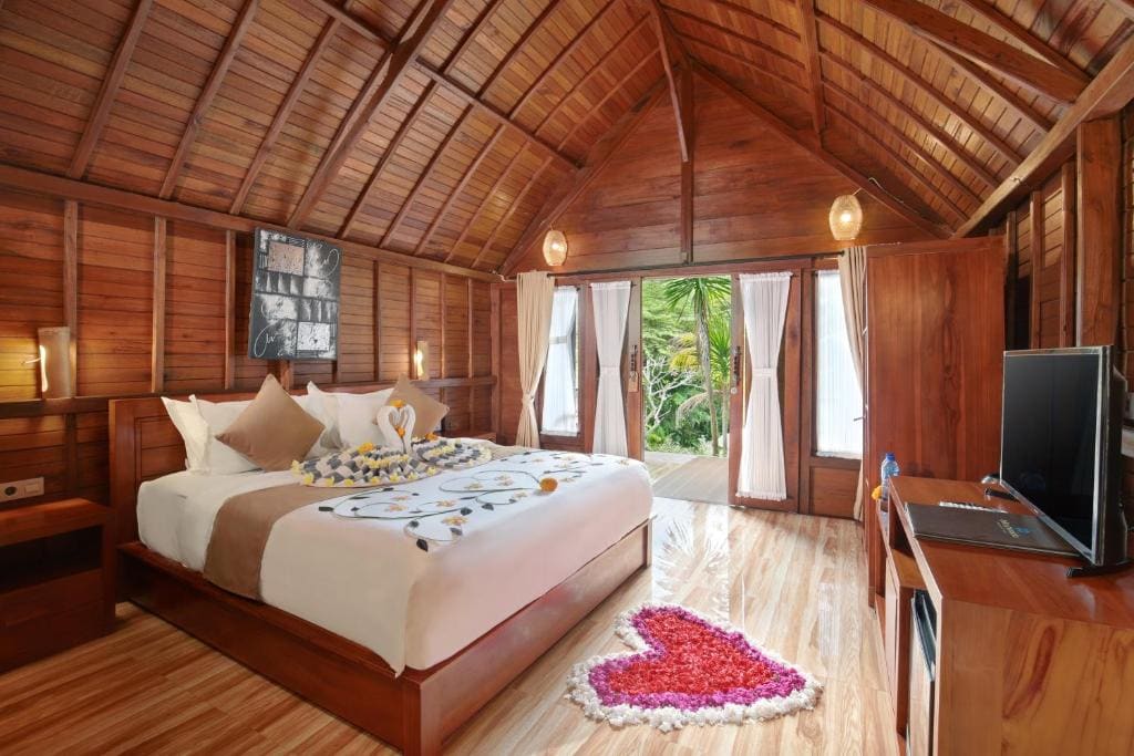 A spacious bedroom features wooden walls and a high ceiling, creating a cozy atmosphere. A queen-sized bed is adorned with decorative elements, including flower arrangements. Large glass doors open to the terrace, allowing natural light and garden views. A flat-screen TV is positioned nearby.
