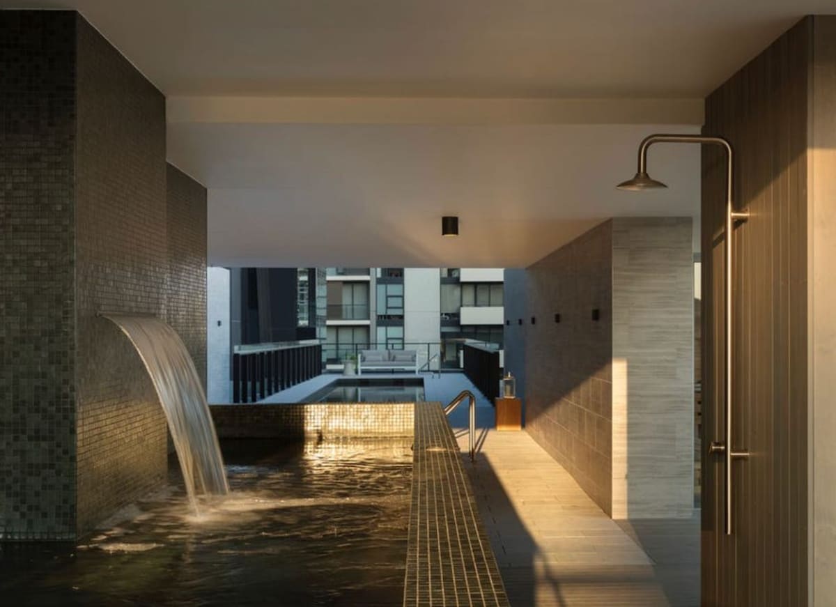 A sleek outdoor pool area is featured, with a waterfall cascading into the water. Dark tiled walls and ample natural light highlight the modern design. A shower is located nearby, enhancing the space's functionality for relaxation and comfort.