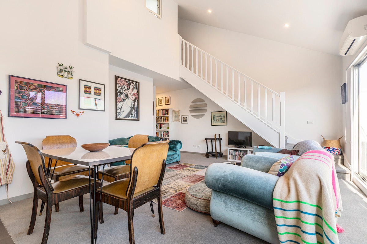 An open-plan living and dining area is highlighted by a dining table for four and a plush sofa draped with a colorful blanket. Artwork adorns the walls, and a staircase leads to the upper level. Natural light enters through large glass doors that open to an outdoor space.