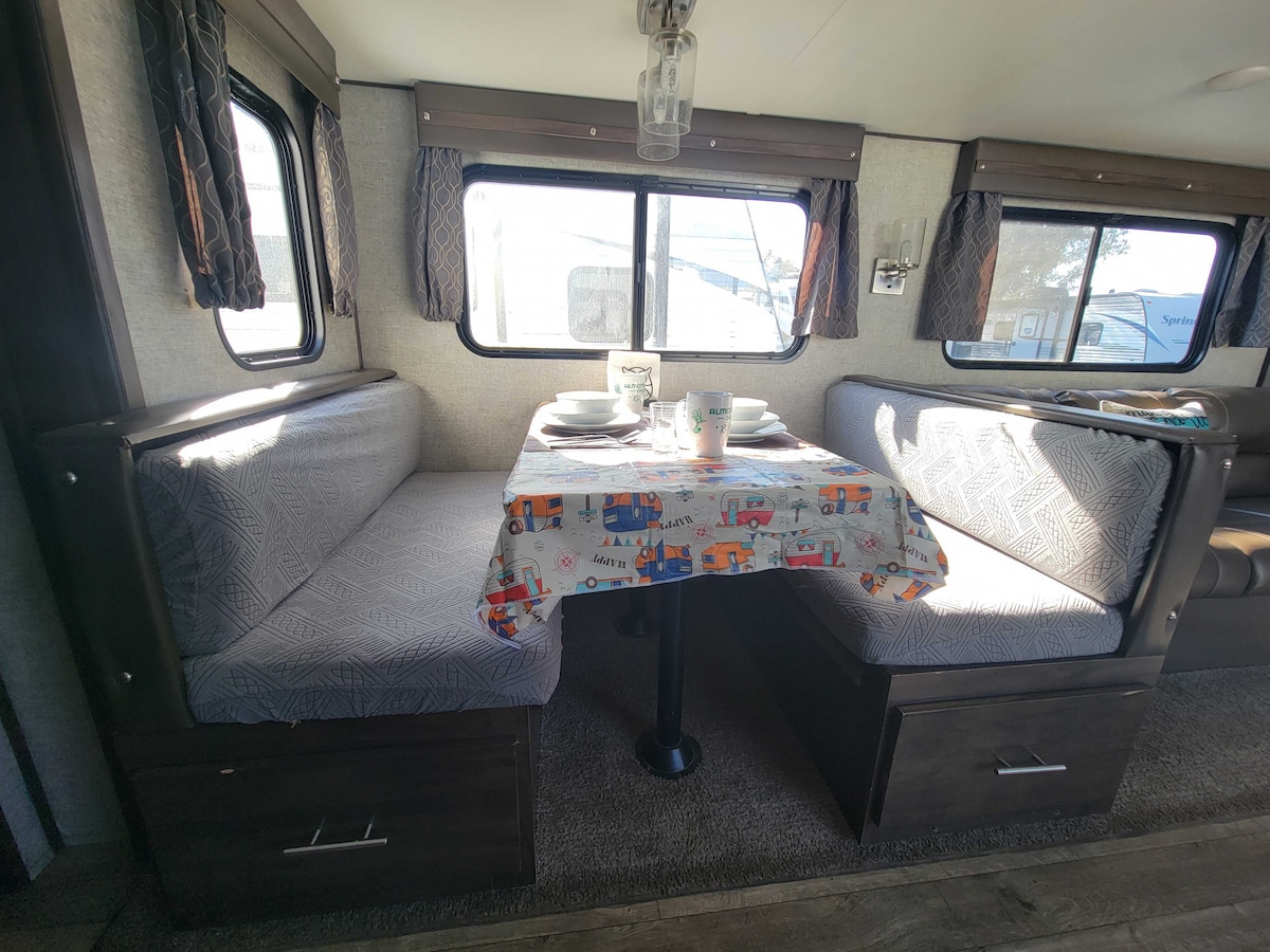 A compact dining area features a table set with dishes and utensils. Booth seating is provided on either side, with patterned tablecloth adding color. Several windows allow natural light to fill the space, creating a bright atmosphere within the travel trailer.