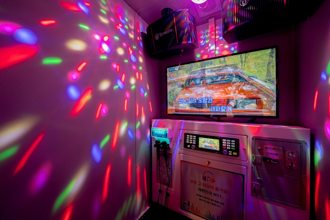 A vibrant karaoke room is adorned with colorful light patterns reflecting off the walls, creating an entertaining ambiance. A large screen displays lyrics for guests, while a control panel is positioned below it. The space is equipped for a playful singing experience.