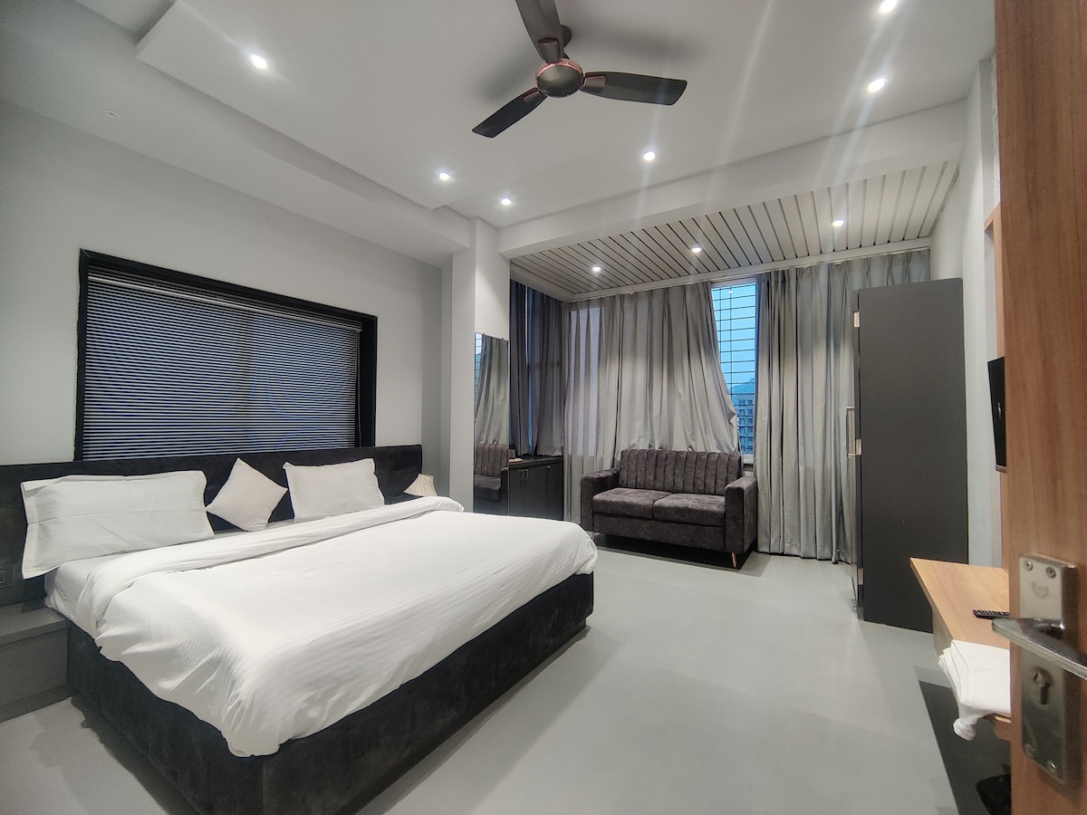 A modern bedroom features a spacious bed with crisp white linens, accompanied by two pillows. A dark gray sofa and a work desk are positioned near large windows, draped with sheer curtains. Soft lighting illuminates the room, creating a welcoming ambiance.