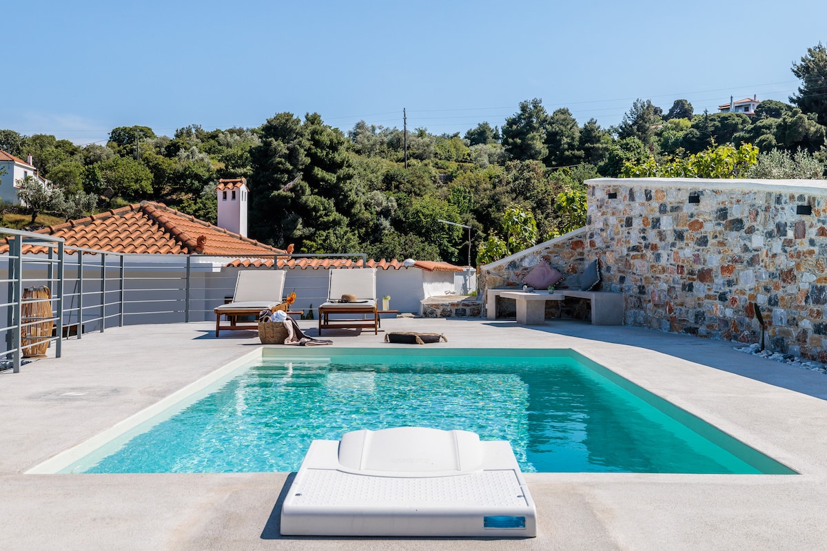 A serene outdoor space features a clear swimming pool surrounded by a smooth patio. Sun loungers are positioned nearby, offering a comfortable resting area. A stone wall adds texture, and the lush greenery in the background provides a natural setting.