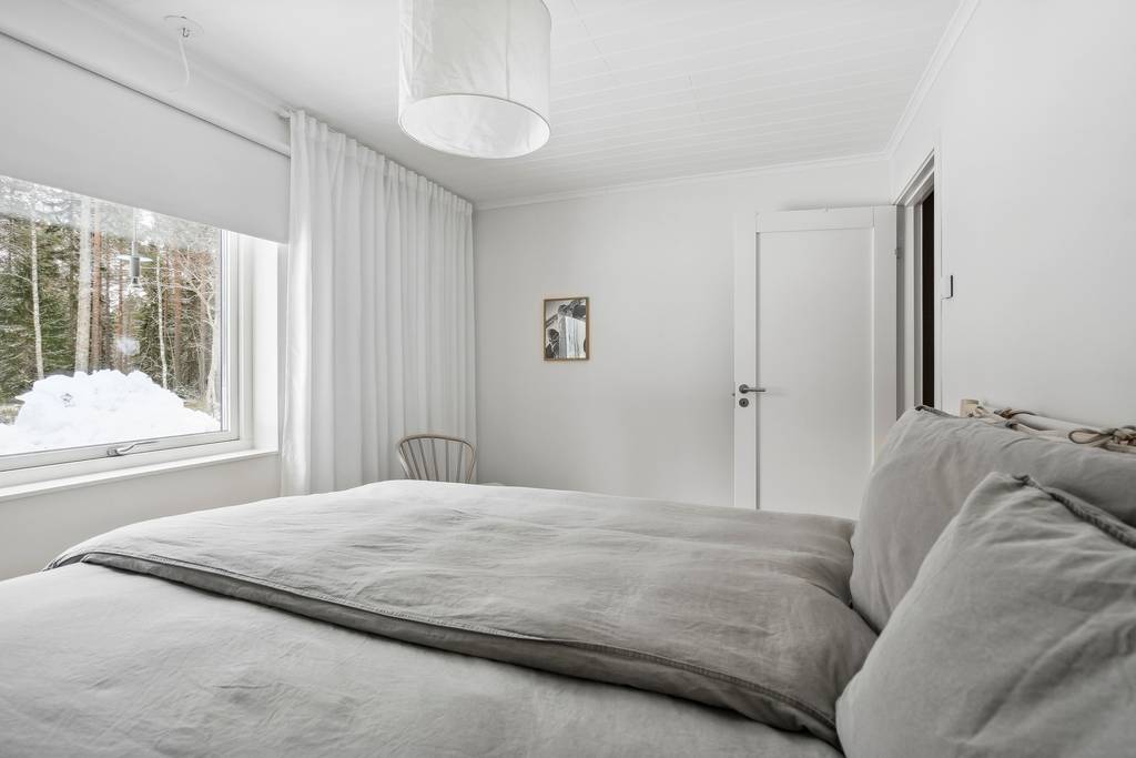 A bright bedroom features a spacious bed with light gray bedding. Soft white curtains frame a window that overlooks a snowy landscape. A simple mirror hangs on the wall, and a wooden chair adds a touch of warmth to the minimalist décor.