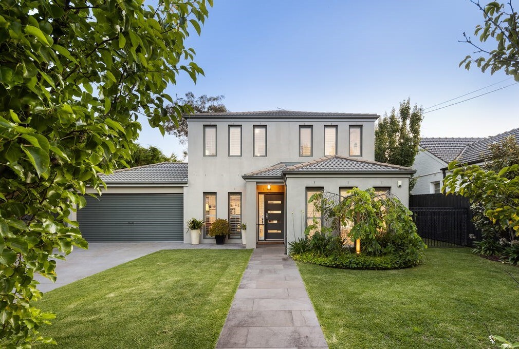 The exterior of the home features a contemporary two-story design, complemented by a well-maintained lawn and landscaped garden. A stone pathway leads to the entrance, flanked by greenery and containers with plants. A garage is visible to the left, enhancing the overall curb appeal.