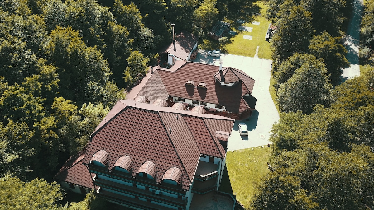 An aerial view captures the guesthouse, showcasing its distinctive roofline and multiple gables. The surrounding greenery of trees and grass creates a natural setting, while a paved area is visible nearby, suitable for various outdoor activities.