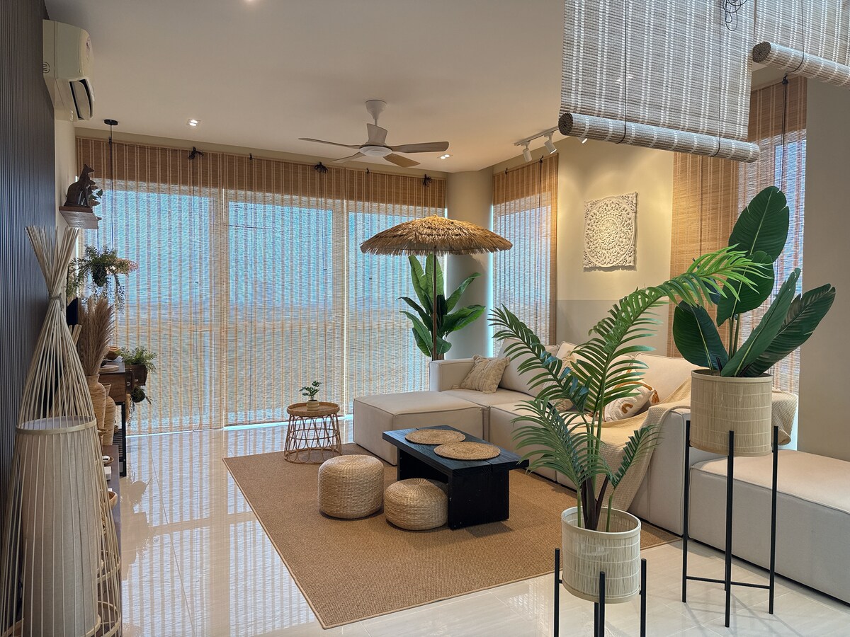 A serene living space is highlighted by large windows allowing natural light. Subtle shades of beige and earthy tones dominate the decor. Stylish furniture, including a sectional sofa, a low wooden table, and potted plants, creates a calming environment, complemented by natural textures and materials.