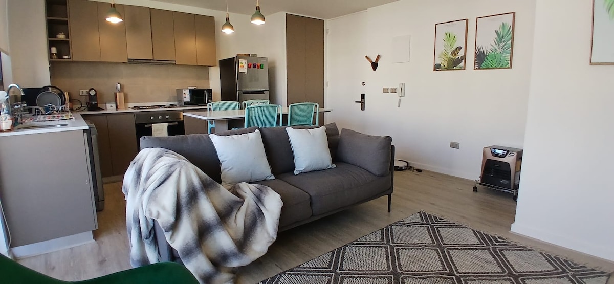 A modern living area is highlighted by a sectional sofa adorned with light-colored cushions and a plaid throw. An open kitchen features stainless steel appliances and a dining table with turquoise chairs. Wall art depicting foliage accents the space, complemented by natural light from large windows.