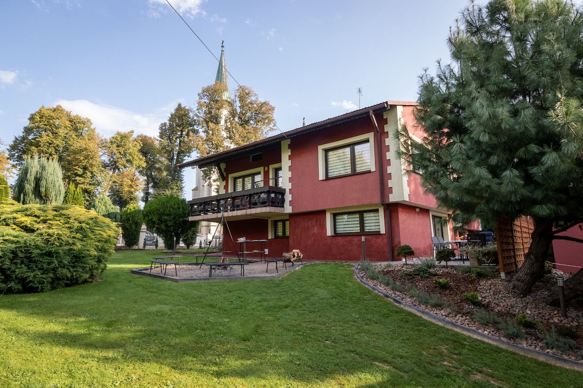 The exterior of the villa showcases a two-story structure with a warm red facade, complemented by balconies on the upper level. Lush green lawns surround the building, with well-kept gardens featuring trees and shrubs. A playground area and a distant church steeple are visible in the background.