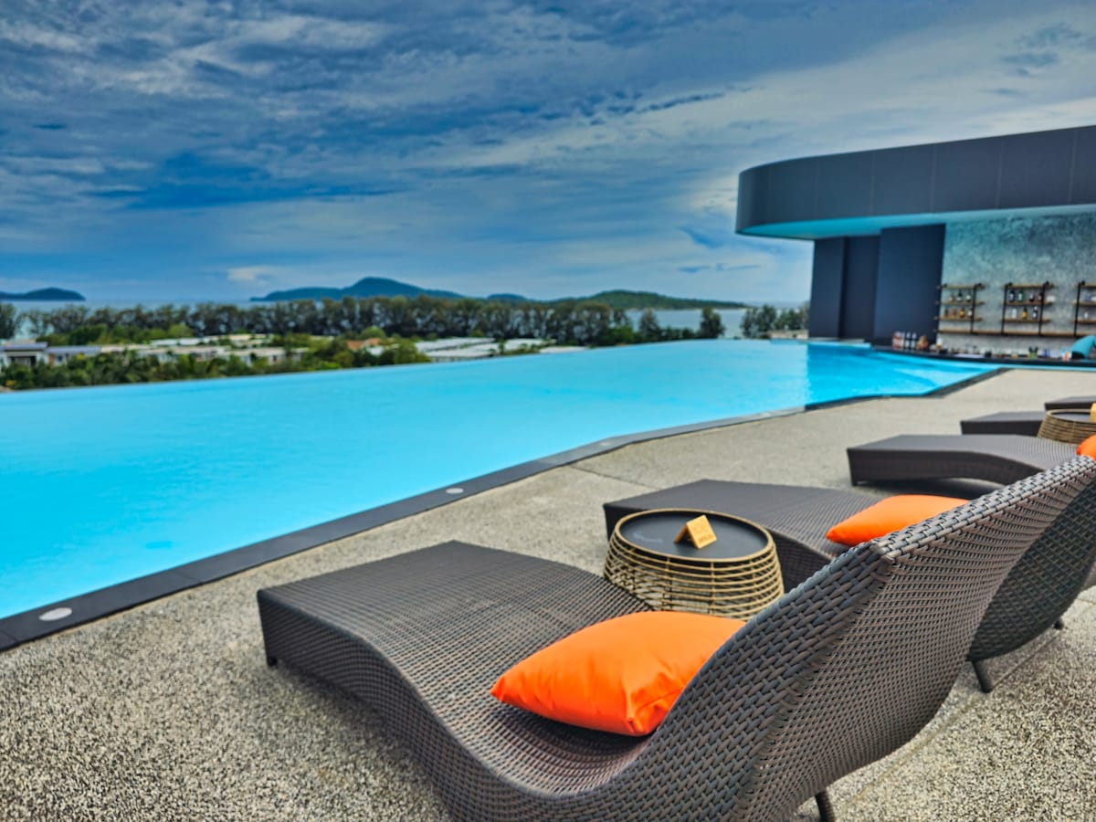 An infinity pool extends towards the horizon, offering captivating views of the surrounding landscape. Sun loungers with bright orange cushions are positioned beside the pool, creating a comfortable space for relaxation. The sleek, modern hotel design is evident in the pool area.