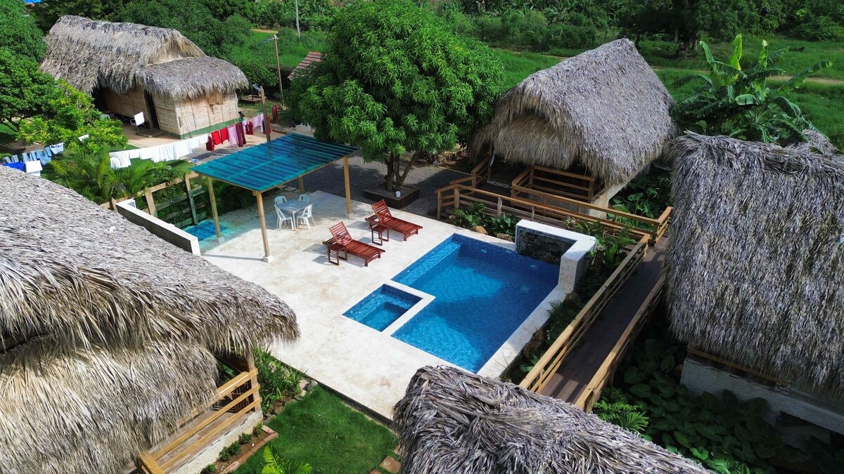 An aerial view reveals several thatched-roof cottages surrounding a pool area. The pool features a children's section and is bordered by sun loungers. Lush greenery is visible in the background, enhancing the natural setting.