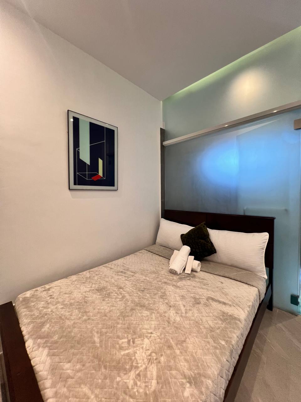 A compact sleeping area is presented, featuring a textured blanket draped over a spacious bed. A decorative pillow complements the bedding. A minimalist piece of wall art adds a touch of color, while a glass partition allows light to filter through from an adjacent area.