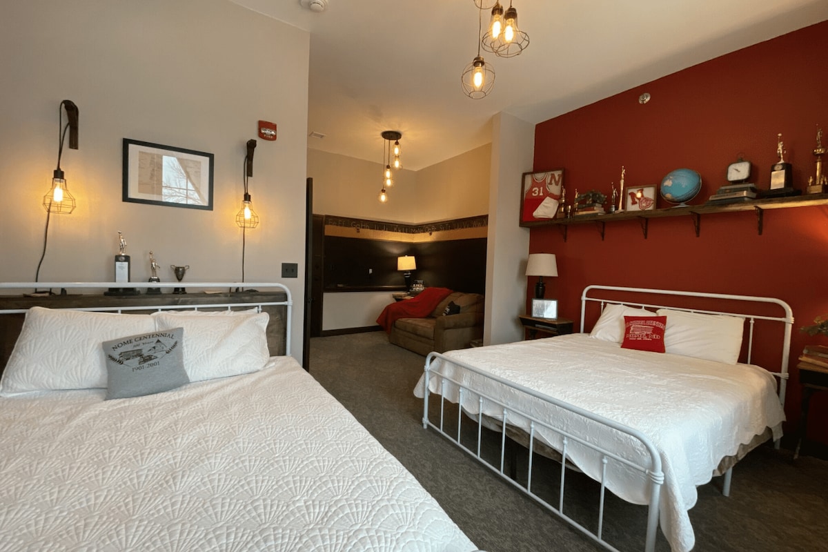The room features two spacious beds with white bedding, framed against a deep red accent wall. A cozy seating area is visible in the background, with warm lighting from hanging fixtures enhancing the ambiance. Decor elements include trophies, pictures, and sports memorabilia.