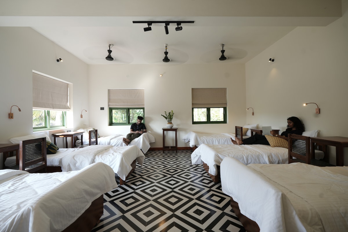 A spacious dormitory features multiple beds with clean white linens arranged symmetrically around the room. Natural light floods in through large windows, while a geometric patterned floor adds a modern touch. Simple wooden nightstands and decorative plants provide a warm ambiance.