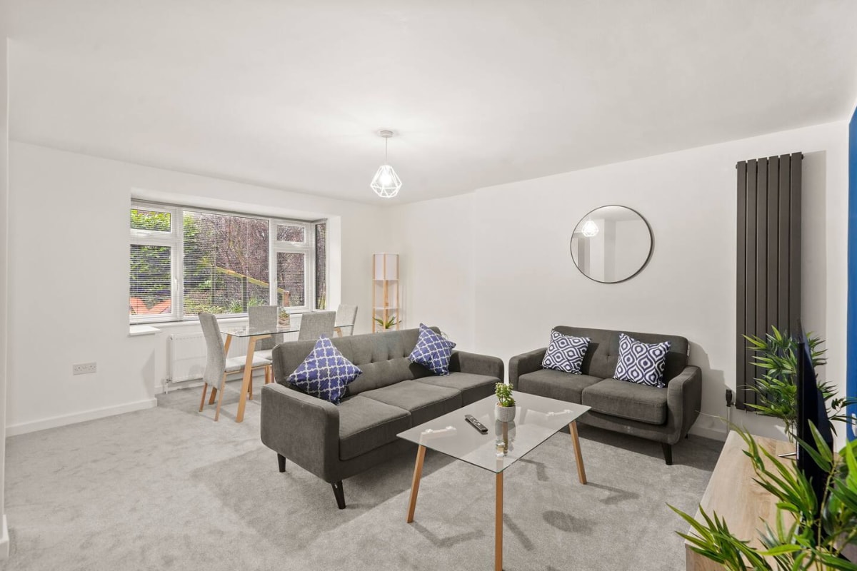 A living area showcases a modern grey sofa adorned with blue-patterned cushions. A glass coffee table sits centrally, complemented by a small decorative plant. In the background, a dining table with chairs is positioned beside a large window, allowing natural light to enter the space.