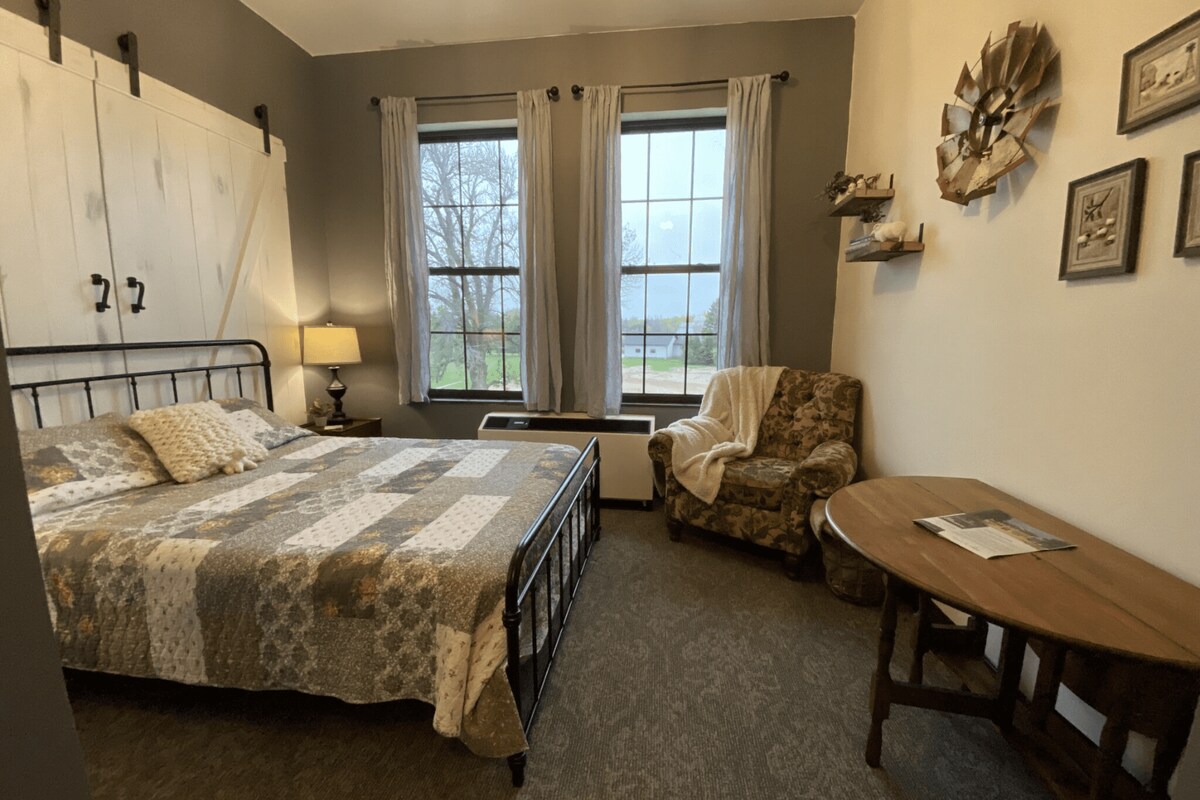 The room features a metal bed with a patchwork quilt and decorative pillows. Natural light enters through two large windows dressed in sheer curtains. A cozy armchair, a round table, and wall décor enhance the space, creating a comfortable atmosphere.