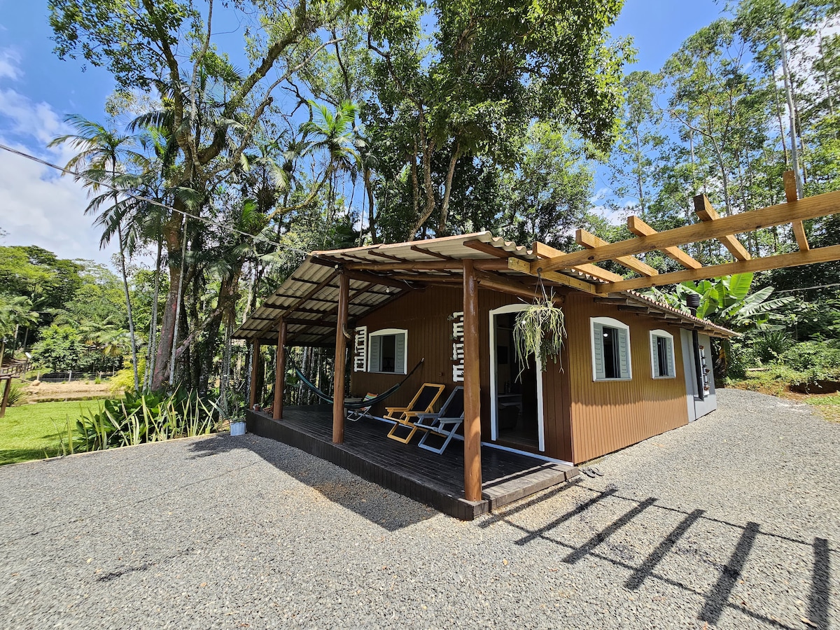A charming wooden house is surrounded by lush greenery and sunlight. The front porch features two lounge chairs and is sheltered by a slatted roof, enhancing the connection to nature. The space invites relaxation amidst the serene landscape.
