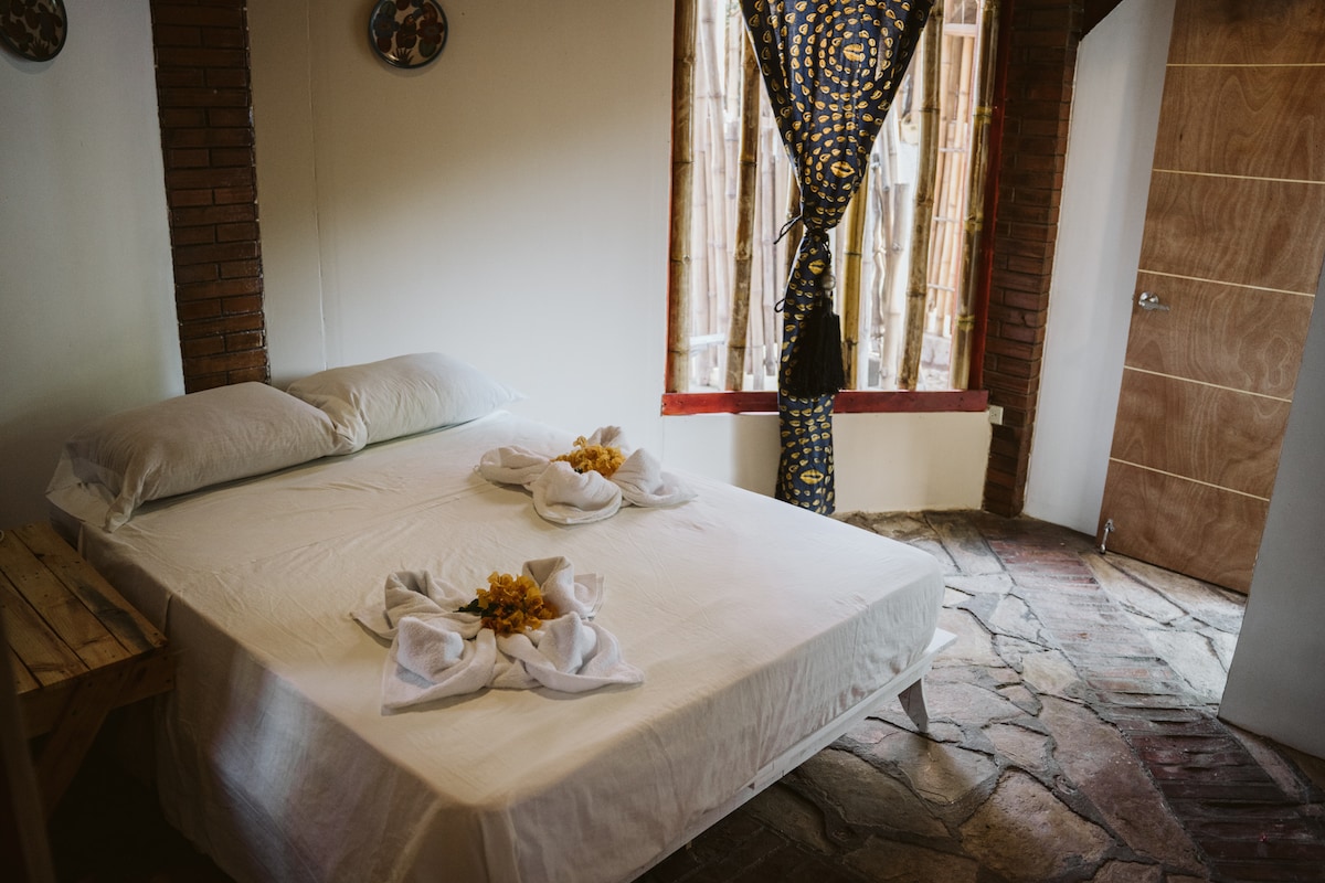 A well-lit room features a spacious bed dressed in white linens, adorned with towel art and floral accents. Exposed brick walls provide a rustic touch, while a patterned curtain allows soft light to filter in. A wooden table is positioned nearby, complementing the earthy stone floor.