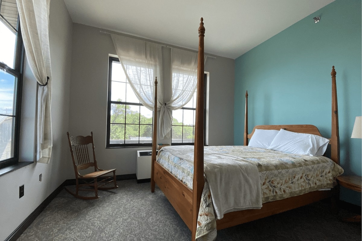 A spacious sleeping area features a wooden four-poster bed dressed with fresh linens. A rocking chair is positioned next to a window, allowing natural light to fill the room. The walls are accented in soft blue, enhancing the open feel of the space.