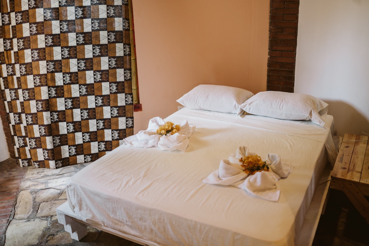 A spacious bed with white linen is neatly arranged, adorned with towel art resembling flowers. The backdrop features a patterned curtain in warm tones, contributing to the room's aesthetic. Exposed brick walls add a rustic touch to the overall ambiance.
